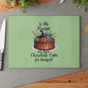 May include: A rectangular glass chopping board with a green background. It features a chocolate cake topped with a witch's hat and flowers. Text on the board reads, "In this House we have Chocolate Cake for breakfast."