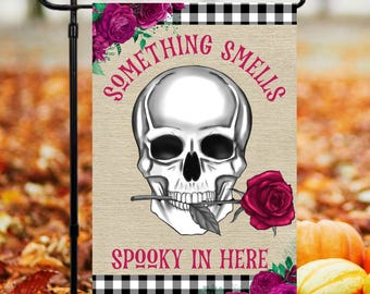 Puede incluir: Una bandera de jardín a cuadros blanco y negro con una calavera blanca sosteniendo una rosa roja. El texto en la bandera dice "Something Smells Spooky In Here".