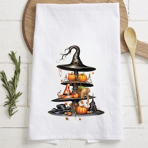 Könnte beinhalten: Weißes Küchentuch mit einem mehrstöckigen Display zum Thema Halloween. Die Präsentation umfasst einen schwarzen Hexenhut, orangefarbene Kürbisse und verschiedene andere Hüte. Das Design ist mit Herbstblättern und dekorativen Elementen versehen.