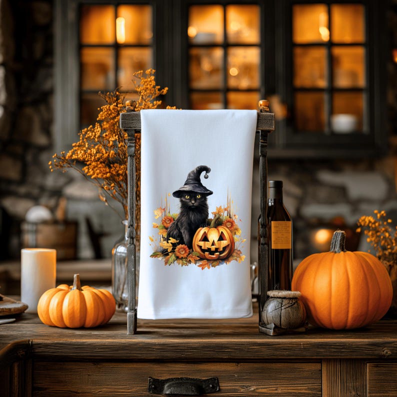 May include: A white kitchen towel with a black cat wearing a witch's hat sitting on a jack-o'-lantern. The cat is surrounded by fall leaves and pumpkins.