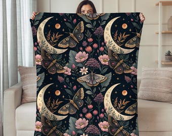May include: A dark blue blanket featuring a repeating pattern of moths, crescent moons, flowers, and stars. The moths are brown and tan, the moons are cream-colored, and the flowers are pink and purple. The blanket is held up by a person.