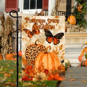 May include: A garden flag with the text "Pumpkin Spice Season" in a brown, orange, and white font. The flag features two butterflies, one with black and orange wings and the other with orange and brown wings. There are also two pumpkins, one with a leopard print design and the other with a traditional orange colour. The flag is surrounded by autumn leaves and flowers.