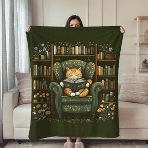 May include: A green throw blanket featuring a whimsical illustration of a cat reading in a green armchair in front of a bookshelf. The cat is orange and white, and the armchair has a floral pattern. The bookshelf is filled with books and flowers.