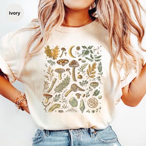 May include: A white t-shirt with a graphic print of mushrooms, leaves, crystals, and other forest elements in shades of brown, green, and gold.