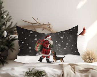 Può includere: Un cuscino rettangolare a tema natalizio. Il cuscino presenta una raffigurazione di Babbo Natale che cammina con un cane bassotto in una scena innevata. Babbo Natale indossa un abito a quadri rossi e porta un sacco di regali. Fiocchi di neve cadono su uno sfondo nero.