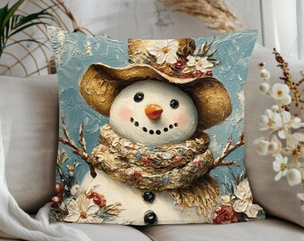 Op de afbeelding: Een decoratief vierkant kussen met een lachende sneeuwman met een bruine hoed versierd met witte bloemen. De sneeuwman heeft een wortelneus, zwarte knoopogen en een sjaal met patroon. De achtergrond is lichtblauw.