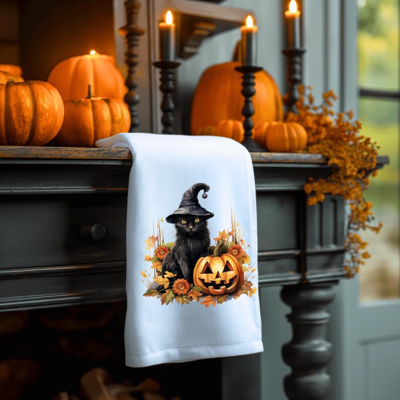 May include: A white kitchen towel with a black cat wearing a witch's hat and holding a jack-o'-lantern. The cat is surrounded by fall leaves and flowers.