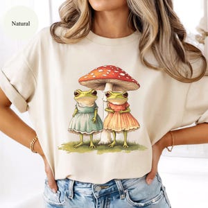 May include: A cream-colored t-shirt featuring a whimsical illustration of two green frogs wearing dresses under a red and white mushroom. The word "Natural" is printed on the shirt.