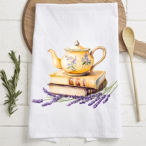 May include: White kitchen towel featuring a watercolor illustration of a yellow teapot with purple floral accents, stacked on top of two books. Lavender sprigs are at the base of the books. A wooden spoon and cutting board are in the background.