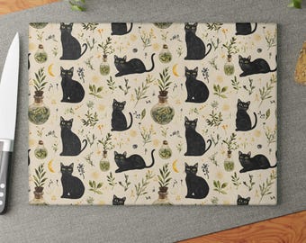 Peut inclure: Planche à découper rectangulaire avec un motif répété de chats noirs, de croissants de lune et d'éléments botaniques sur fond crème. Le motif comprend de petites bouteilles en verre avec du contenu vert et des brins d'herbes. Un couteau argenté est à gauche.