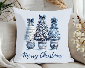 Puede incluir: Un cojín cuadrado blanco con una ilustración en azul y blanco de tres árboles de Navidad en macetas, cada uno con un lazo azul. El árbol central tiene detalles florales y el texto Merry Christmas aparece escrito en azul debajo.
