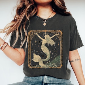 May include: A dark grey t-shirt featuring a vintage illustration of a mermaid within a gold frame against a starry backdrop. The text "Mon Océan" is visible on the right sleeve.