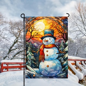 May include: A decorative garden flag featuring a snowman in a winter scene. The snowman has a black top hat, orange scarf, and twig arms. The background shows a sunset with trees and snow-covered ground. The flag is displayed on a black metal pole.