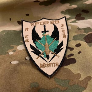 May include: A tan and black embroidered patch with the text "KFOR 28 A Co. Misfits" and a green and brown stylized map with a sword and crossed sticks.