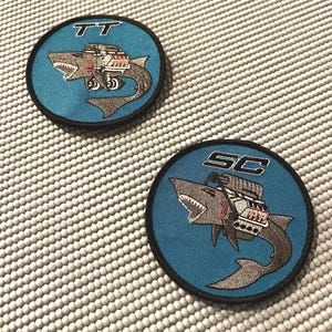 May include: Two round embroidered patches with a blue background and a gray shark with a car engine on its back. The top patch has the letters "TT" in white on the top and the bottom patch has the letters "SC" in white on the top.