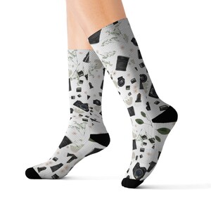 May include: A pair of white socks with a black and white pattern of cameras and flowers. The socks are shown on a model's legs.