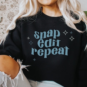 May include: Black sweatshirt with the text "snap edit repeat" in blue lettering with small white stars around the text.