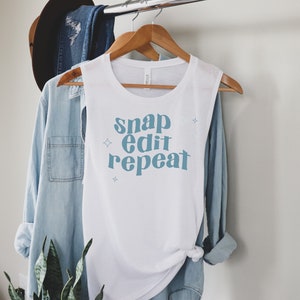 May include: White sleeveless shirt with the text "snap edit repeat" in blue lettering with small white stars.