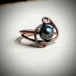 May include: A copper wire ring with a black hematite stone in the center. The ring is made of two loops of wire that are twisted together.