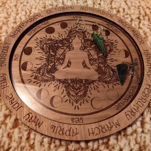 May include: A wooden pendulum board with engraved months, phrases, and a central Buddha figure. Two green crystal pendulums hang from chains. The board features the text "yes," "no," and "maybe."