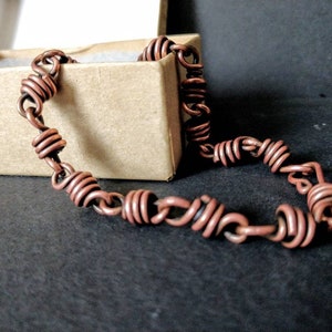 May include: A copper wire bracelet with a coiled design. The bracelet is lying on a black surface and is in a small box.