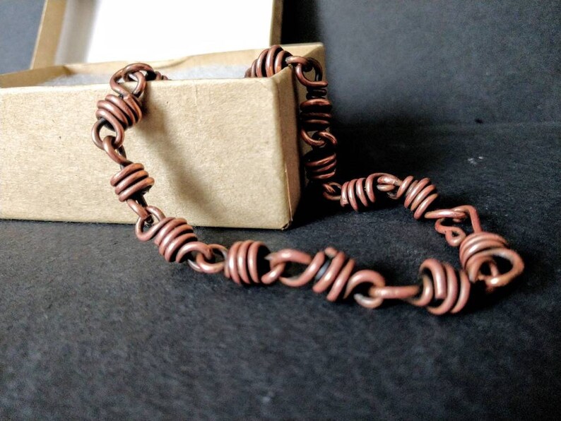May include: A copper wire bracelet with a coiled design. The bracelet is lying on a black surface and is in a small box.