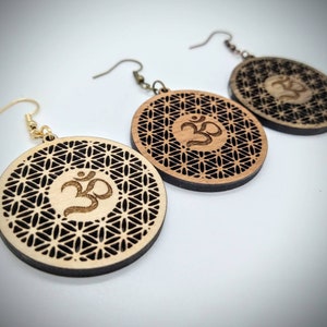 May include: Three pairs of wooden earrings with a laser-cut design featuring the Om symbol and a flower of life pattern. The earrings are brown and have gold hooks.