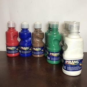 Könnte beinhalten: Eine Sammlung von Prang Temperafarben-Flaschen in verschiedenen Farben, darunter Rot, Blau, Braun, Grün und Weiß. Jede Flasche hat einen weißen Deckel und ein Etikett mit dem Prang-Logo und Text. Die Flaschen stehen auf einer dunkelbraunen Oberfläche.