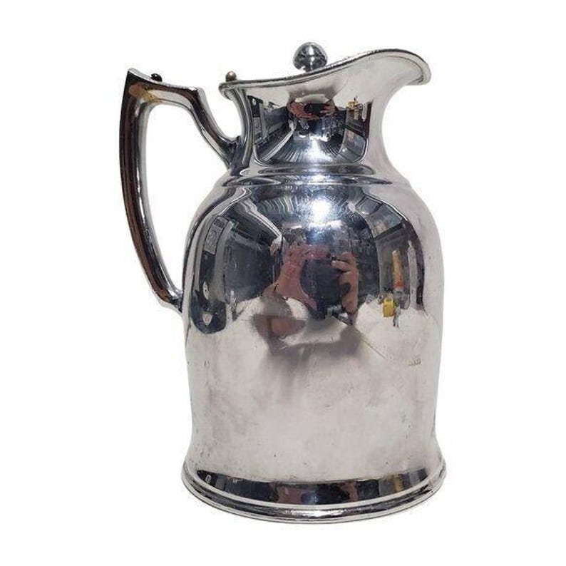 K&ouml;nnte beinhalten: Eine gl&auml;nzende, silberfarbene Kanne mit gebogenem Griff und Deckel. Die Kanne hat einen abgerundeten K&ouml;rper und einen Ausguss zum Ausgie&szlig;en. Die Oberfl&auml;che reflektiert die Umgebung. Die Kanne ist wahrscheinlich aus Metall.