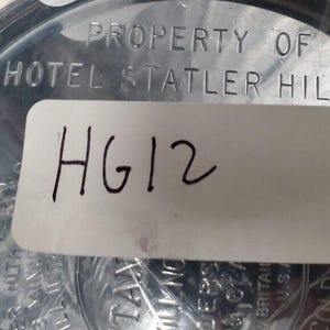 K&ouml;nnte beinhalten: Nahaufnahme eines silberfarbenen Objekts mit dem gepr&auml;gten Text "PROPERTY OF HOTEL STATLER HILL". Ein wei&szlig;er Aufkleber mit dem handgeschriebenen Text "HG12" ist auf dem Objekt angebracht. Das Objekt scheint ein Sammlerst&uuml;ck oder Souvenir zu sein.