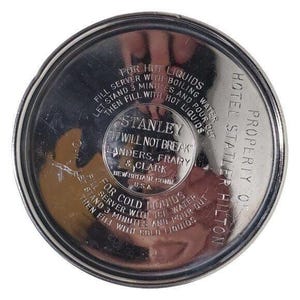 K&ouml;nnte beinhalten: Ein kreisf&ouml;rmiger Stanley-Thermoskannen-Deckel aus Edelstahl mit gepr&auml;gtem Text. Der Text enth&auml;lt Anweisungen f&uuml;r hei&szlig;e und kalte Fl&uuml;ssigkeiten, das Stanley-Logo und "Property of Hotel Statler Hilton."