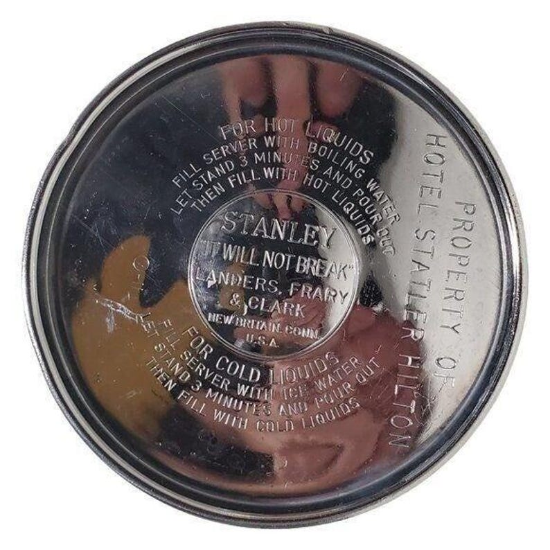 K&ouml;nnte beinhalten: Ein kreisf&ouml;rmiger Stanley-Thermoskannen-Deckel aus Edelstahl mit gepr&auml;gtem Text. Der Text enth&auml;lt Anweisungen f&uuml;r hei&szlig;e und kalte Fl&uuml;ssigkeiten, das Stanley-Logo und "Property of Hotel Statler Hilton."