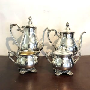 Puede incluir: Un juego de té plateado que incluye una tetera, una lechera, un azucarero y una tetera más pequeña. El juego tiene asas ornamentadas y bases decorativas. La tetera tiene un pico curvo y una tapa con un adorno decorativo.