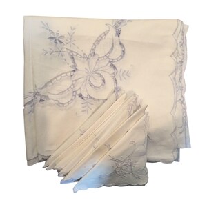 May include: A white tablecloth and a set of matching napkins featuring intricate light blue floral embroidery. The fabric has a scalloped edge with decorative stitching. The embroidery displays detailed leaf and petal patterns on the corners.