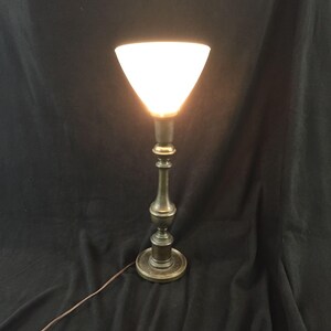 May include: An antique brass table lamp with a white, inverted conical shade. The lamp has a detailed, ornate stem and a round base. The lamp is lit, casting a warm glow. A brown electrical cord extends from the base.