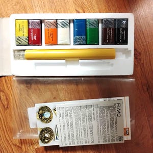 May include: A FIMO polymer clay set with various colored clay blocks, a yellow roller, and instructions. The clay colors include yellow, red, orange, white, blue, green, brown, and black. The set is in a white box with a clear plastic cover.