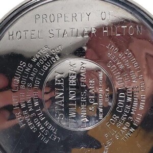 K&ouml;nnte beinhalten: Nahaufnahme eines silbernen Stanley Thermosdeckels. Der Deckel tr&auml;gt gepr&auml;gten Text, darunter "PROPERTY OF HOTEL STATLER HILTON" und Anweisungen f&uuml;r hei&szlig;e und kalte Fl&uuml;ssigkeiten. Der Text enth&auml;lt auch "STANLEY" und "IT WILL NOT BREAK."