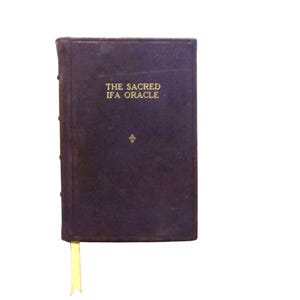 Op de afbeelding: Een donkerpaars boek met gouden letters met de tekst "THE SACRED IFA ORACLE". Een gouden bladwijzer is zichtbaar. Onder de titel staat een klein gouden embleem. Het boek heeft een gestructureerde kaft.