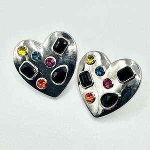 May include: A pair of heart-shaped earrings with a silver finish. Each earring is adorned with a variety of colorful gemstones, including black, yellow, blue, pink, and orange. The gemstones are set in a geometric pattern.