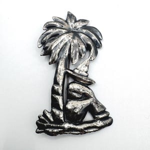 May include: A silver-toned metal brooch depicting a person sitting under a palm tree. The person is wearing a hat. The brooch has a detailed design and is set against a white background.