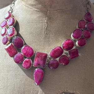 Può includere: Una collana di tendenza con una cascata di grandi pietre di rubino sfaccettate in varie forme e dimensioni. La collana è realizzata in metallo argentato e presenta un ciondolo a forma di lacrima in rubino.