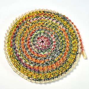 May include: A colorful circular mat made from recycled materials. The mat is made of a variety of colors, including red, orange, yellow, green, and blue. The mat is woven in a spiral pattern.