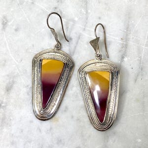 Puede incluir: Un par de pendientes plateados con forma de escudo. Cada pendiente presenta una piedra central con un degradado de colores, que va del amarillo al burdeos oscuro. Los bordes están detallados con un patrón texturizado y los ganchos son sencillos.