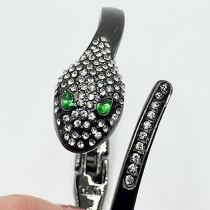 May include: Black snake-shaped bracelet with a hinged design. The snake's head is encrusted with clear rhinestones and features two green gemstone eyes. The bracelet's body is also adorned with rhinestones.