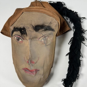 Könnte beinhalten: Eine Vintage-Stoffmaske mit bemaltem Gesicht, braunem Stoffhut und einem langen schwarzen Quasten.