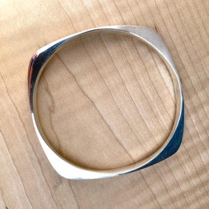 Puede incluir: Un brazalete cuadrado plateado con esquinas redondeadas. El brazalete tiene una superficie pulida y reflectante. La joya se exhibe sobre una superficie de madera de color marrón claro. El brazalete es de diseño moderno y minimalista.