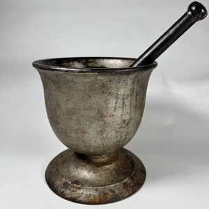 May include: A vintage metal mortar and pestle set. The mortar is a silver-toned bowl with a wide rim and a pedestal base. A black pestle rests inside the bowl. The set has a rustic, aged appearance, suggesting it is antique.