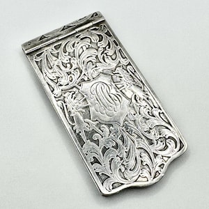 May include: A silver-coloured antique case with detailed floral and scrollwork engraving. The case has a central shield with a monogram and is hinged at the top. The design is highly detailed and symmetrical, typical of the period.