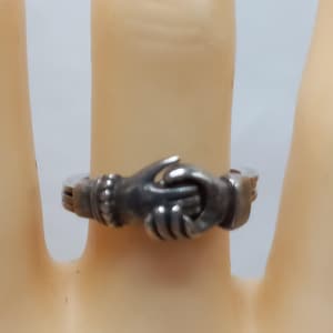 May include: A silver ring featuring a detailed design of two clasped hands. The ring has a vintage or antique appearance, with intricate detailing on the hands and band. The ring is shown on a finger.