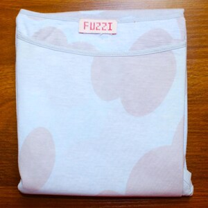 May include: Folded light blue t-shirt with large pink cloud-like shapes. The shirt has a light gray neckline and a small rectangular tag with the word "FUZZI" in pink letters.